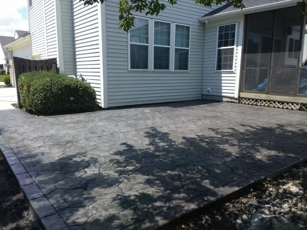A large stamped concrete patio in an irregular ashlar stone pattern, running along the back of a pale blue sided house with a screened porch.