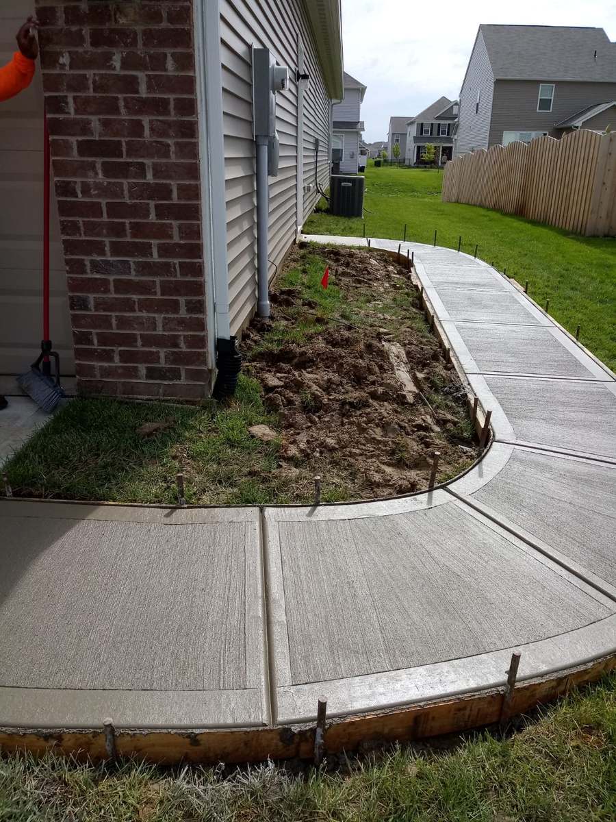A curved concrete sidewalk freshly poured beside a brick house corner, with wooden forms and steel stakes still in place along the curve and bare soil behind it.
